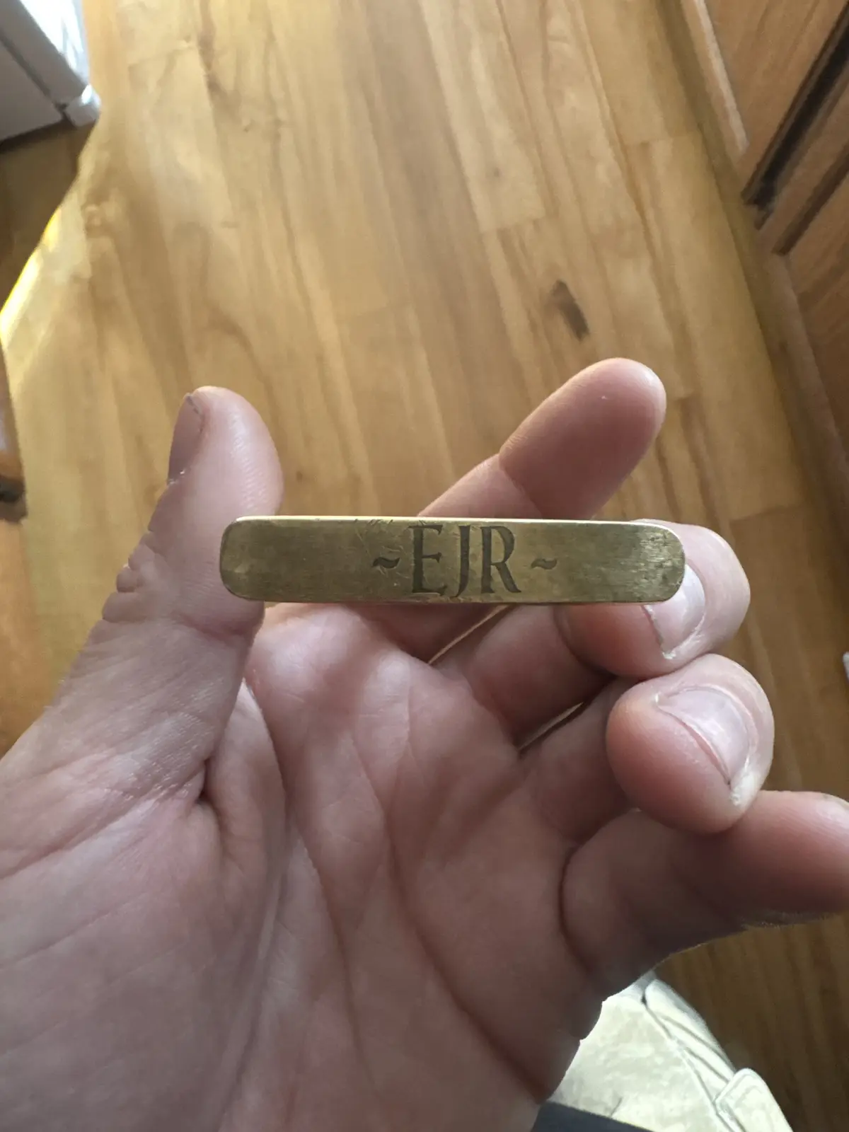 A hand holds a small, brass-colored metal bar engraved with the letters '~EJR~' against a wooden floor background. The text…