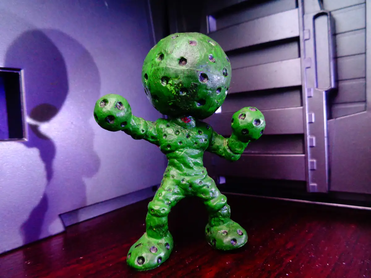 A green, textured figurine resembling a humanoid with a round head and limbs, covered in numerous holes, stands on a dark…