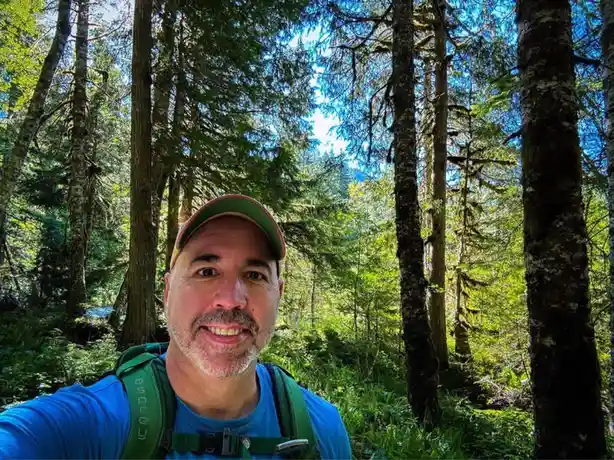 A man with a beard and cap, wearing a blue shirt and green backpack straps, takes a selfie in a sunlit forest with tall…