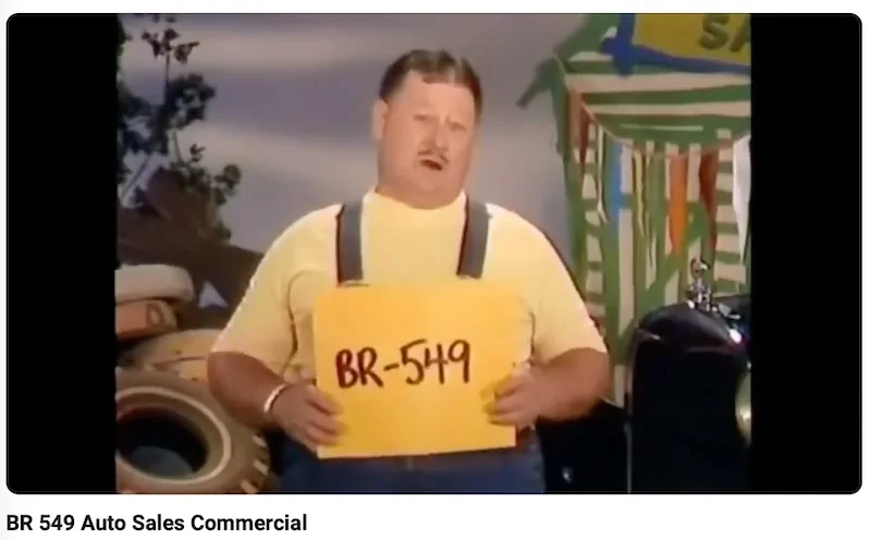 A man in a yellow shirt and overalls holds a yellow sign with 'BR-549' written on it. He stands in front of a backdrop with…