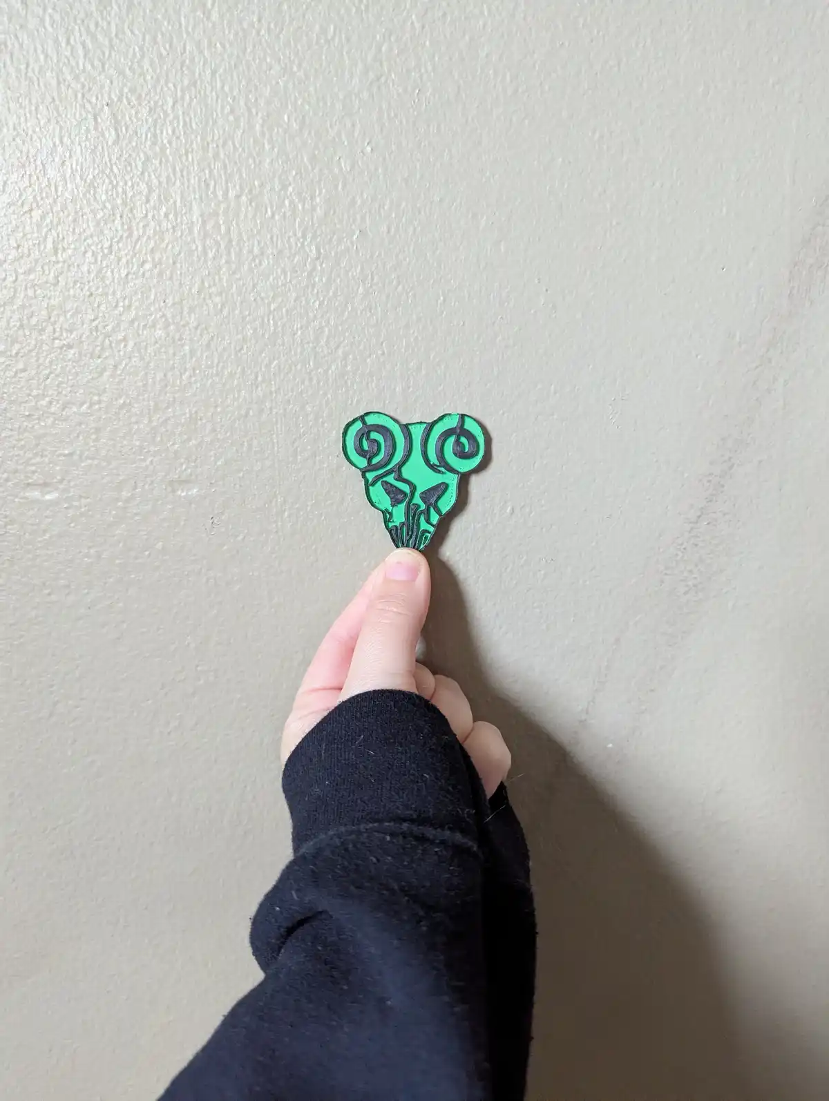 A hand holds a small green pin shaped like a stylized skull with spiral horns against a plain beige wall. The pin is being…