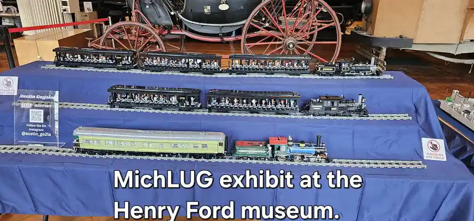Three rows of detailed LEGO train models are displayed on a blue cloth-covered table in a museum, with vintage carriages…