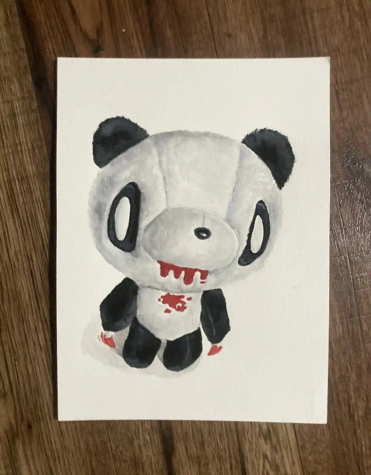 A watercolor drawing of a chibi-style panda plushie on white paper, sitting on a wooden surface. The panda has large black…