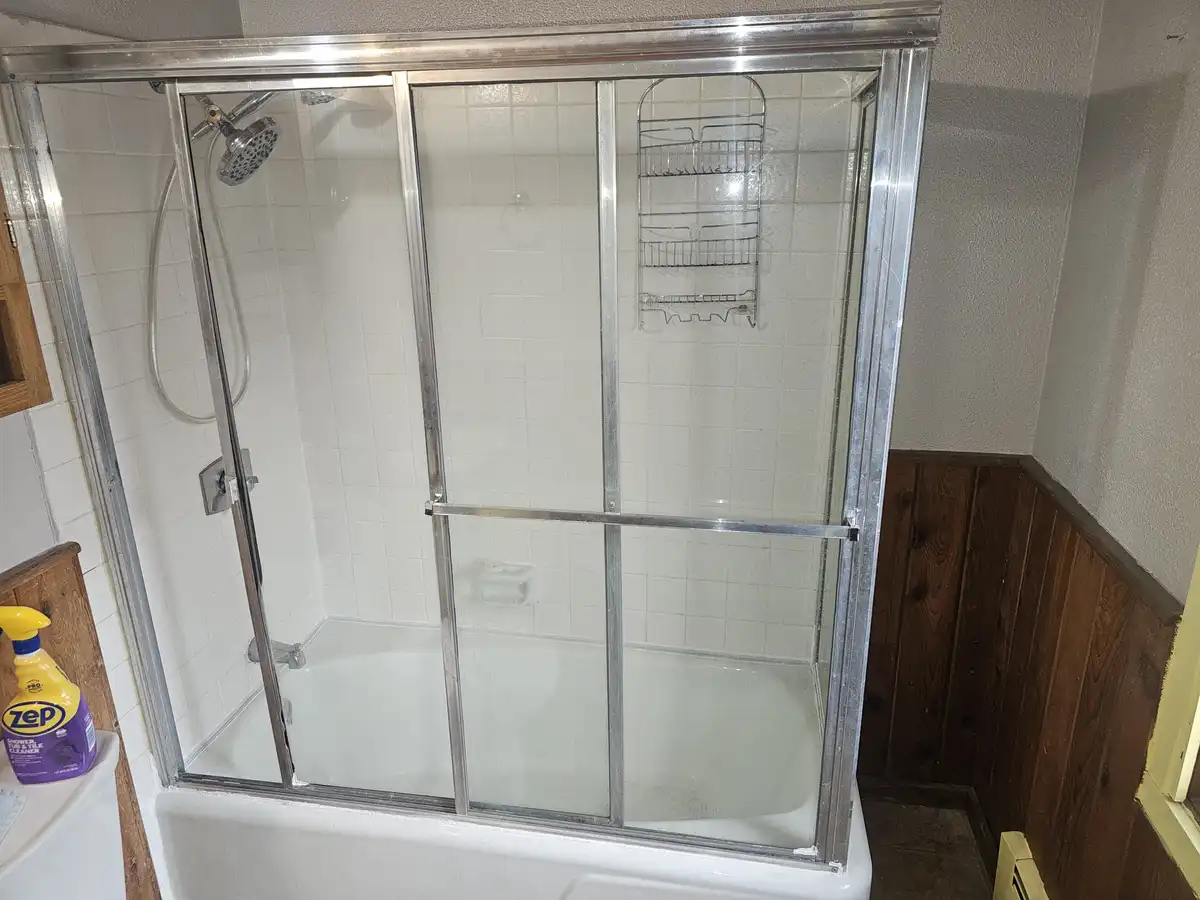 A clean, empty shower stall with a chrome-framed sliding glass door, white tiled walls, and a metal caddy mounted inside. A…
