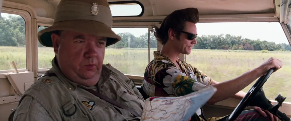Two men are in a vehicle; the driver, wearing sunglasses and a colorful shirt, smiles while steering, while the passenger…