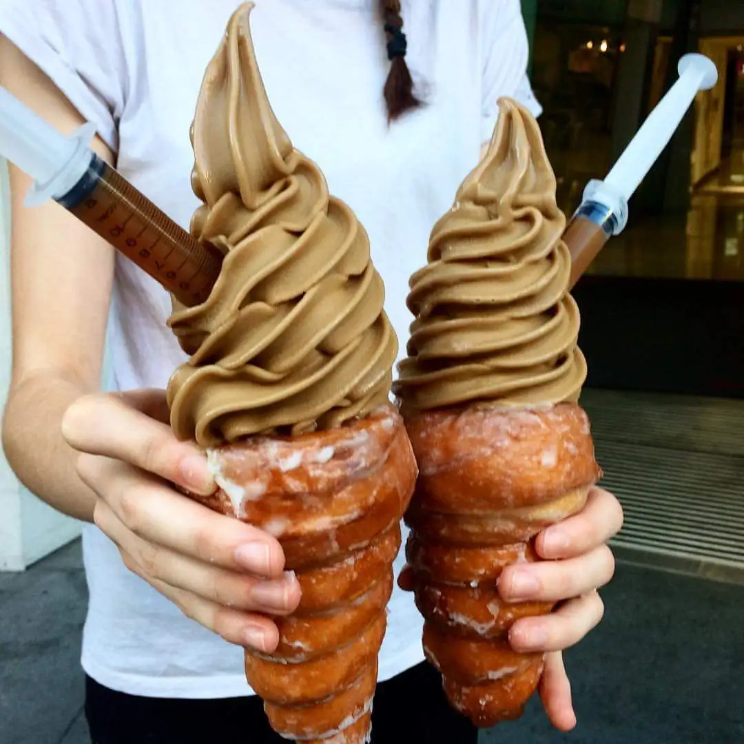 A person holds two giant, cone-shaped desserts made of stacked donuts with swirls of brown soft-serve ice cream on top. Each…