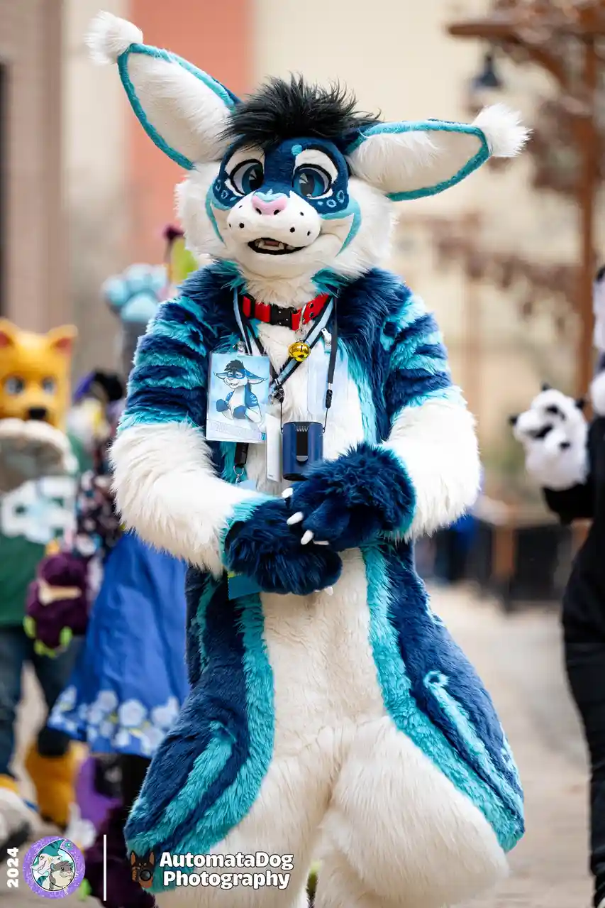 A person in a detailed blue and white fursuit resembling a fox or canine character stands in a crowd at an event, wearing a…