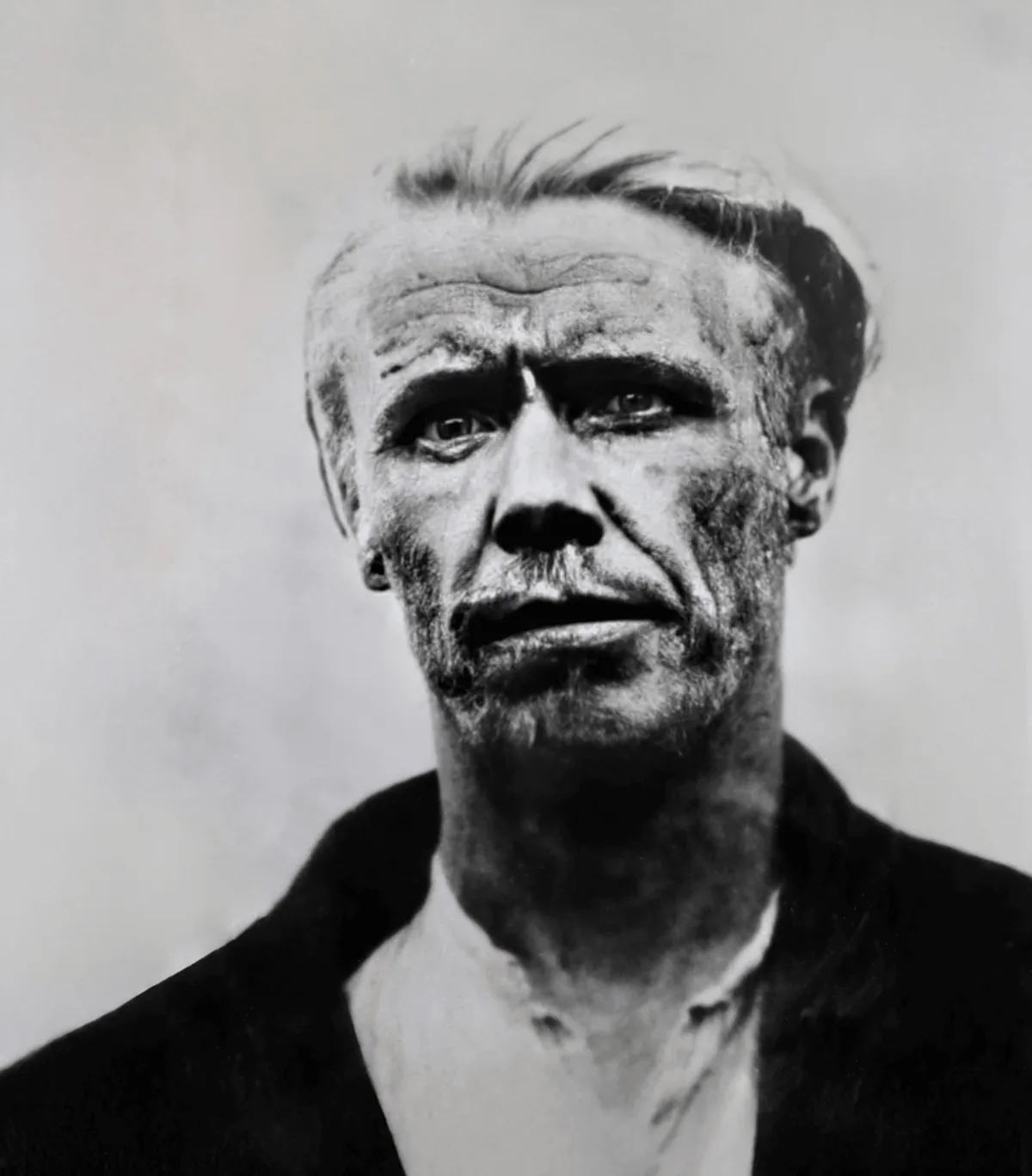 A black-and-white portrait of a man with a weathered, intense expression, looking directly at the camera. His face is…