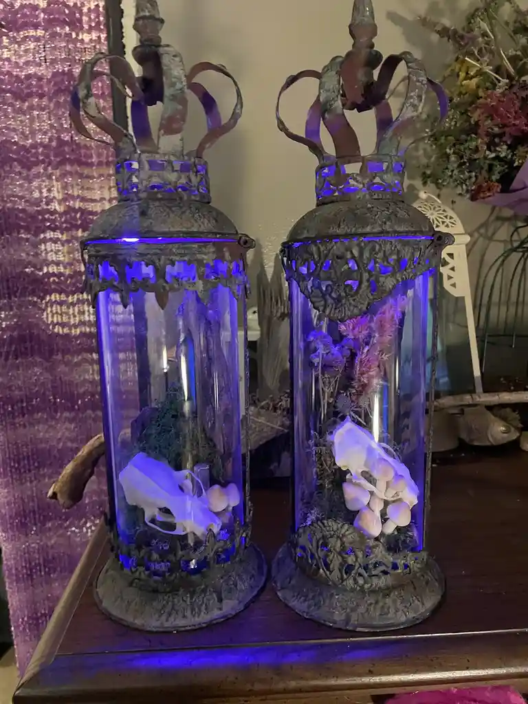 Two ornate, crown-topped glass lanterns with blue LED lighting sit side by side on a wooden surface. Each lantern contains a…