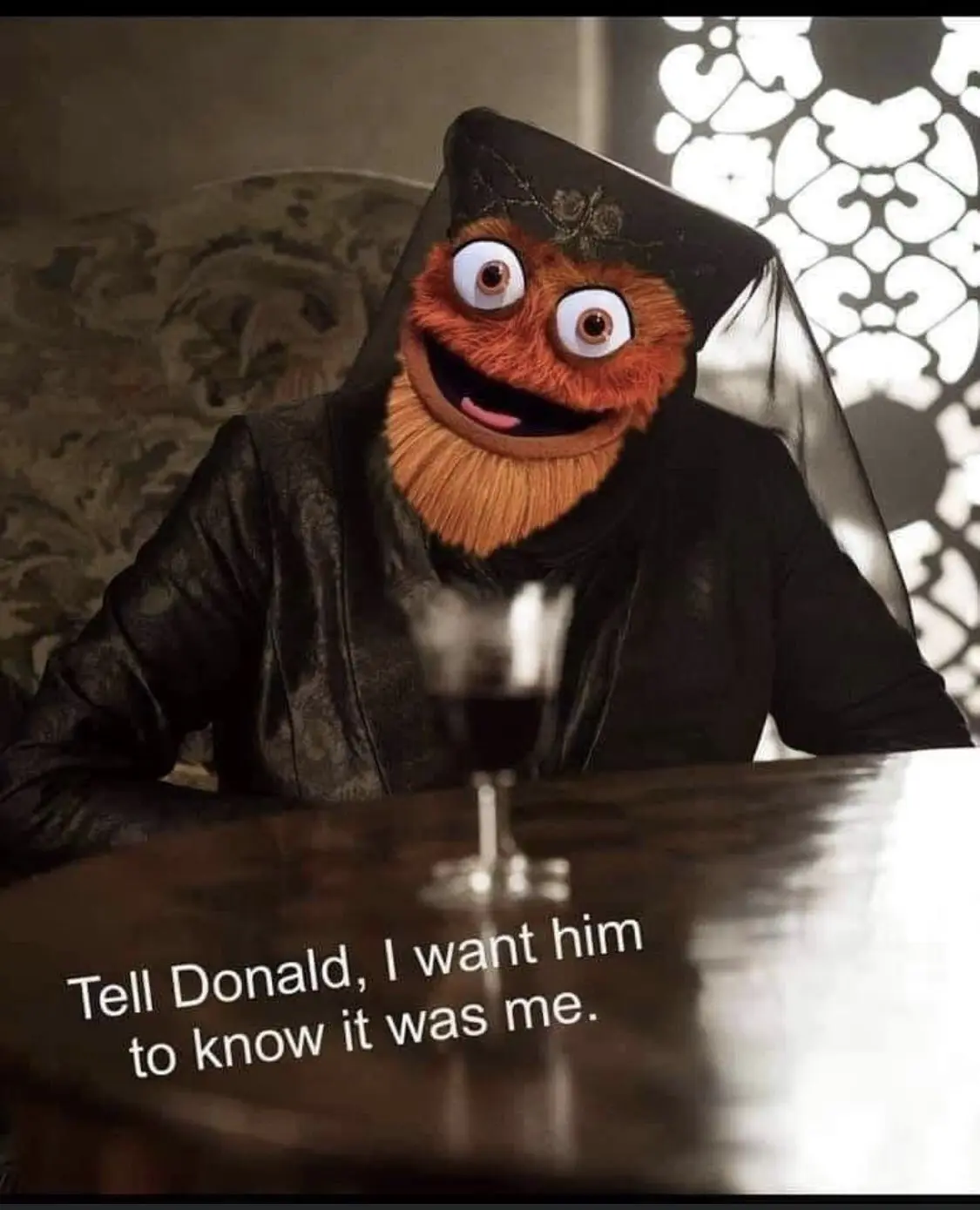 An orange, furry puppet with large eyes and a beard, dressed in a black hooded robe and veil, sits at a dark wooden table…