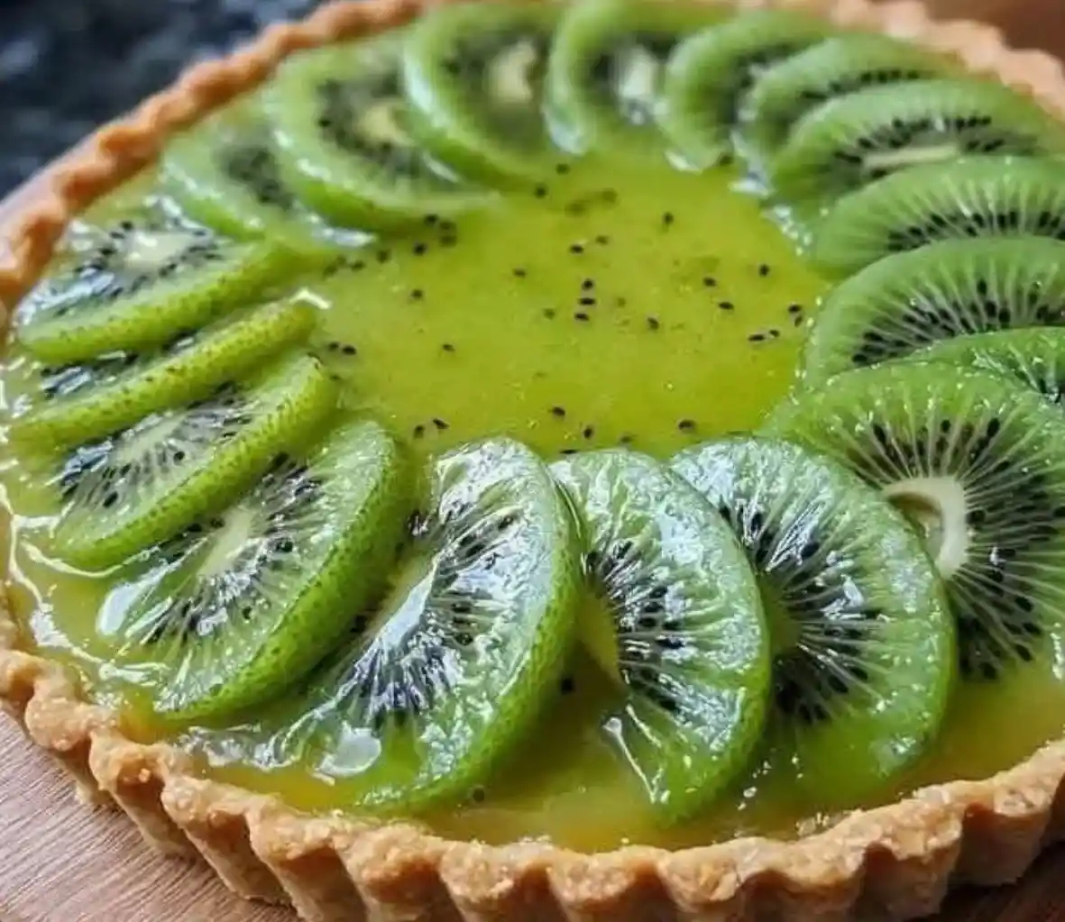 A close-up photo of a golden-brown tart crust filled with a vibrant green kiwi custard and topped with neatly arranged…