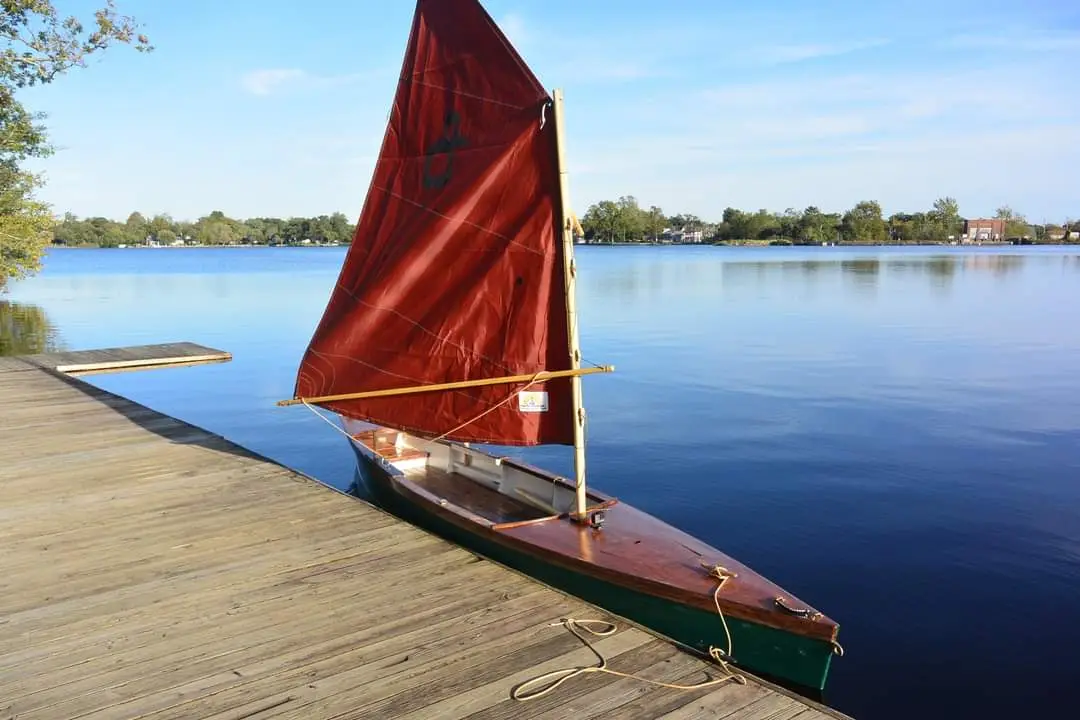 A small wooden sailboat with a red sail is docked at a wooden pier on a calm lake under a clear blue sky. The sail has a…