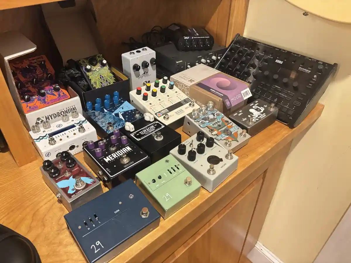 A wooden shelf is cluttered with numerous guitar effect pedals of various colors, sizes, and designs, including brands like…