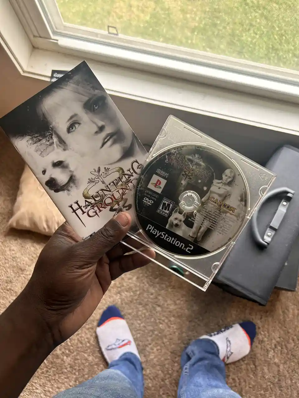 A person holds a PlayStation 2 game case and disc for 'Haunting Ground,' featuring a woman and a white dog on the cover. The…