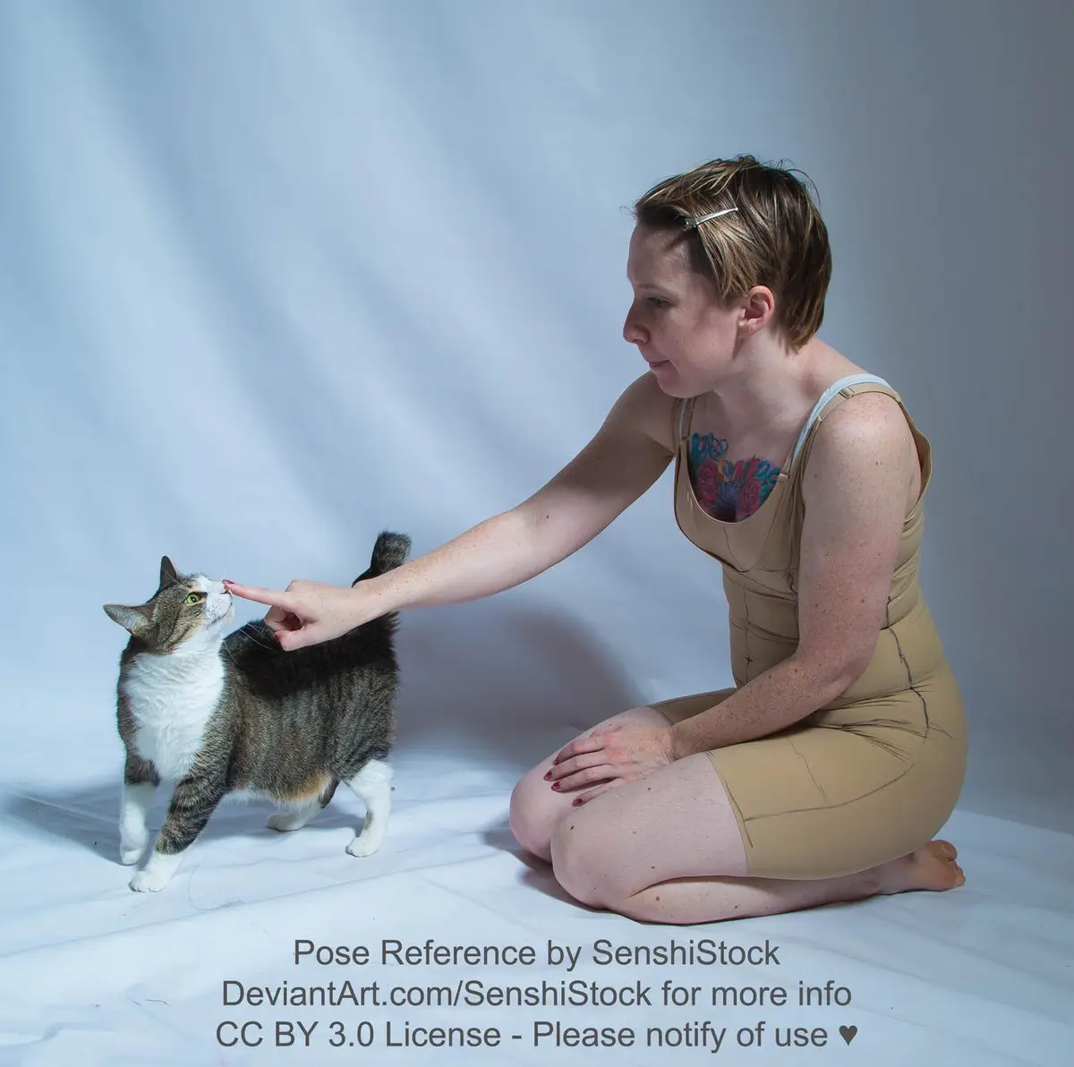 A woman in a beige form-fitting bodysuit with visible seam lines kneels on a white studio floor, gently touching the nose of…