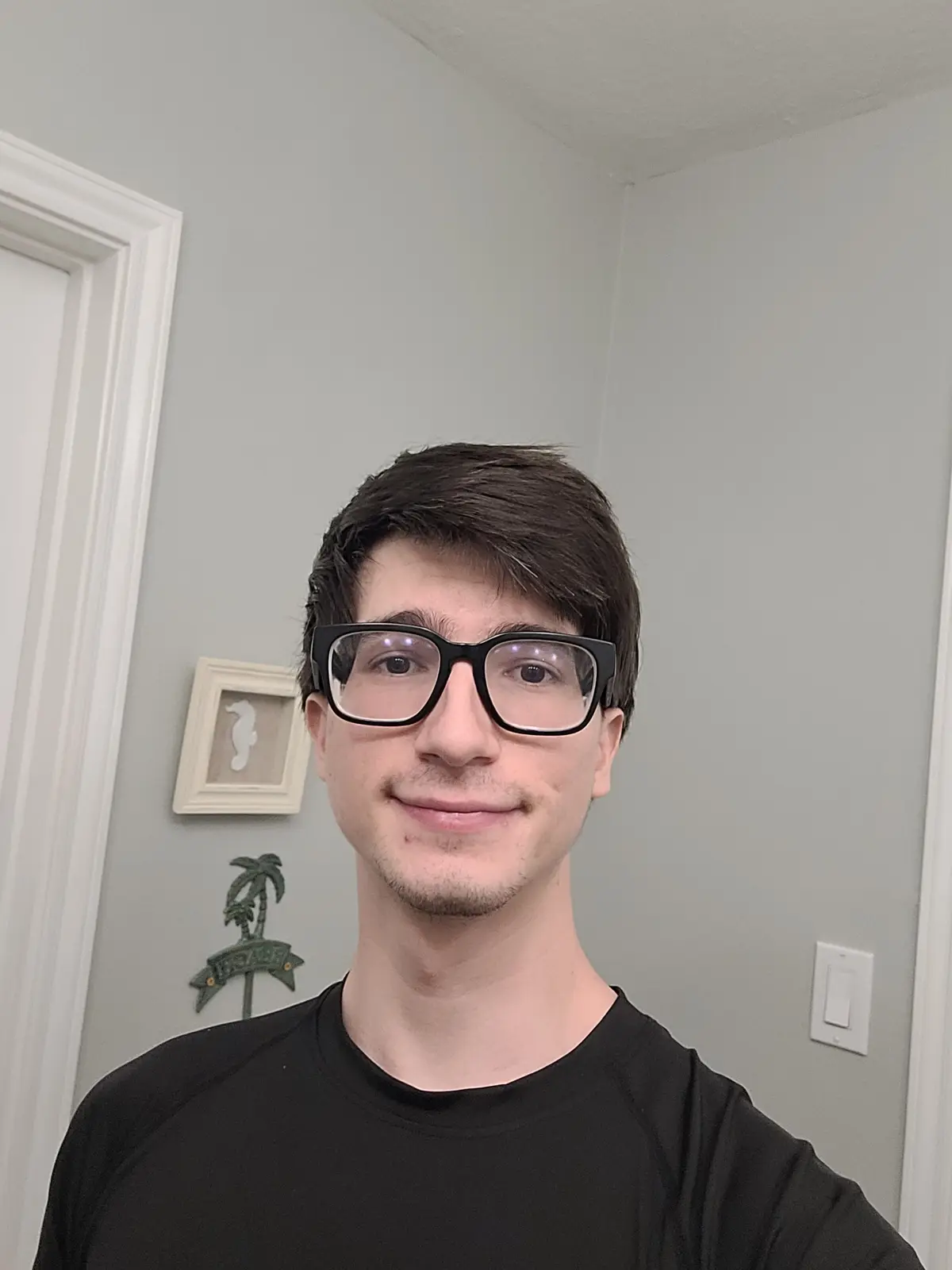 A young man with dark hair and black-rimmed glasses smiles at the camera while wearing a black t-shirt, standing in a room…
