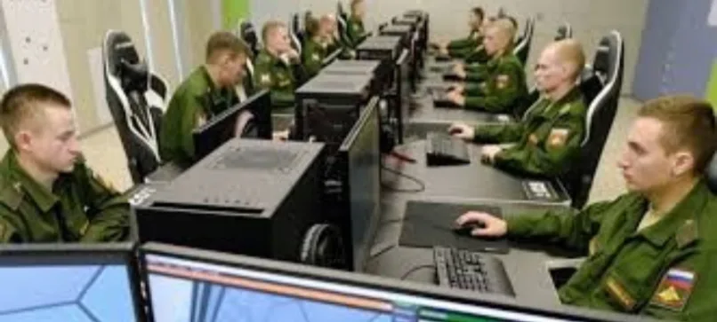 a row of men in green military uniforms sit at desks with large computer monitors and gaming-style chairs, focused on their…