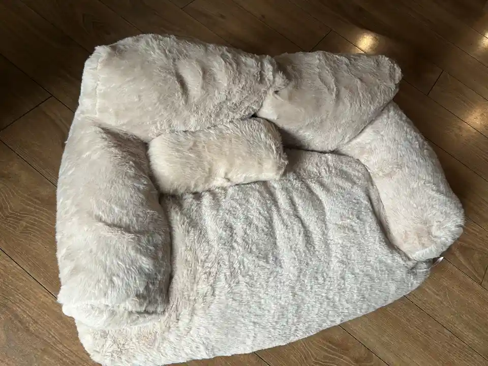 A fluffy, beige pet bed shaped like a cozy cave is photographed from above on a wooden floor. The bed is unoccupied and…