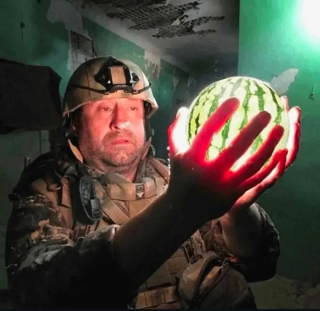 A man in military gear, wearing a helmet with a headlamp, holds a watermelon in a dilapidated, green-walled room. His hand…