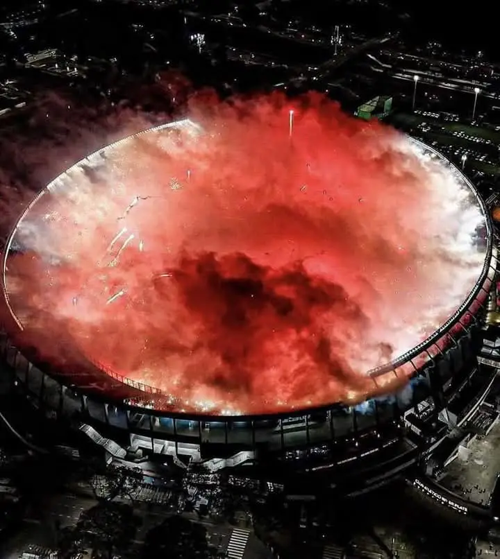 An aerial night photo shows a large circular stadium engulfed in thick red smoke and flames, with bright orange fire visible…
