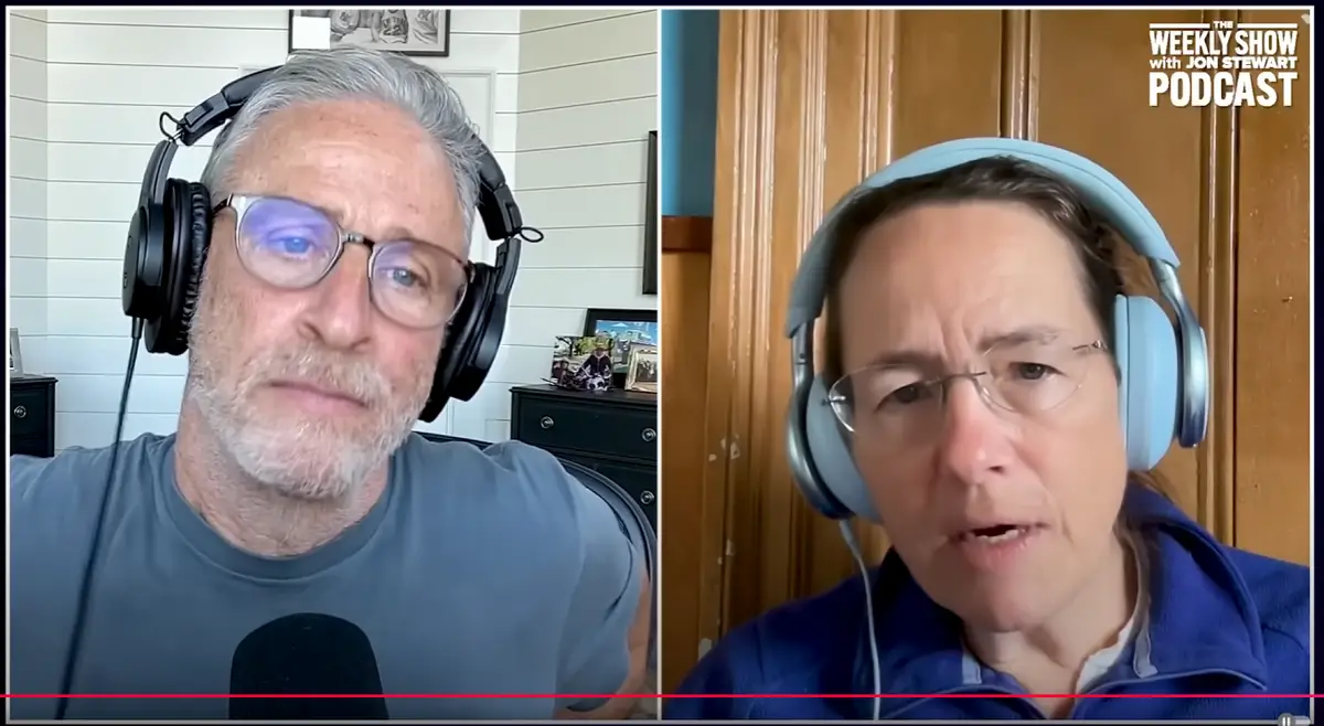 A split-screen video call shows Jon Stewart on the left wearing black headphones and a gray t-shirt, looking serious, and a…