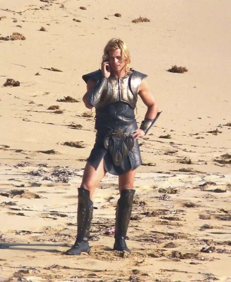 A man in ancient-style armor and tall boots stands on a sandy beach holding a modern cell phone to his ear, looking slightly…