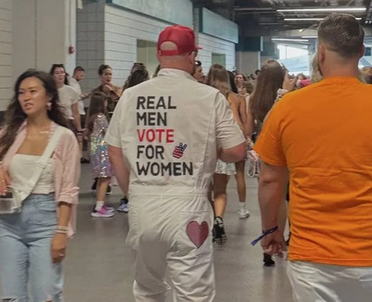 A man in a white jumpsuit with 'REAL MEN VOTE FOR WOMEN' printed on the back and a heart on the rear pocket walks through a…