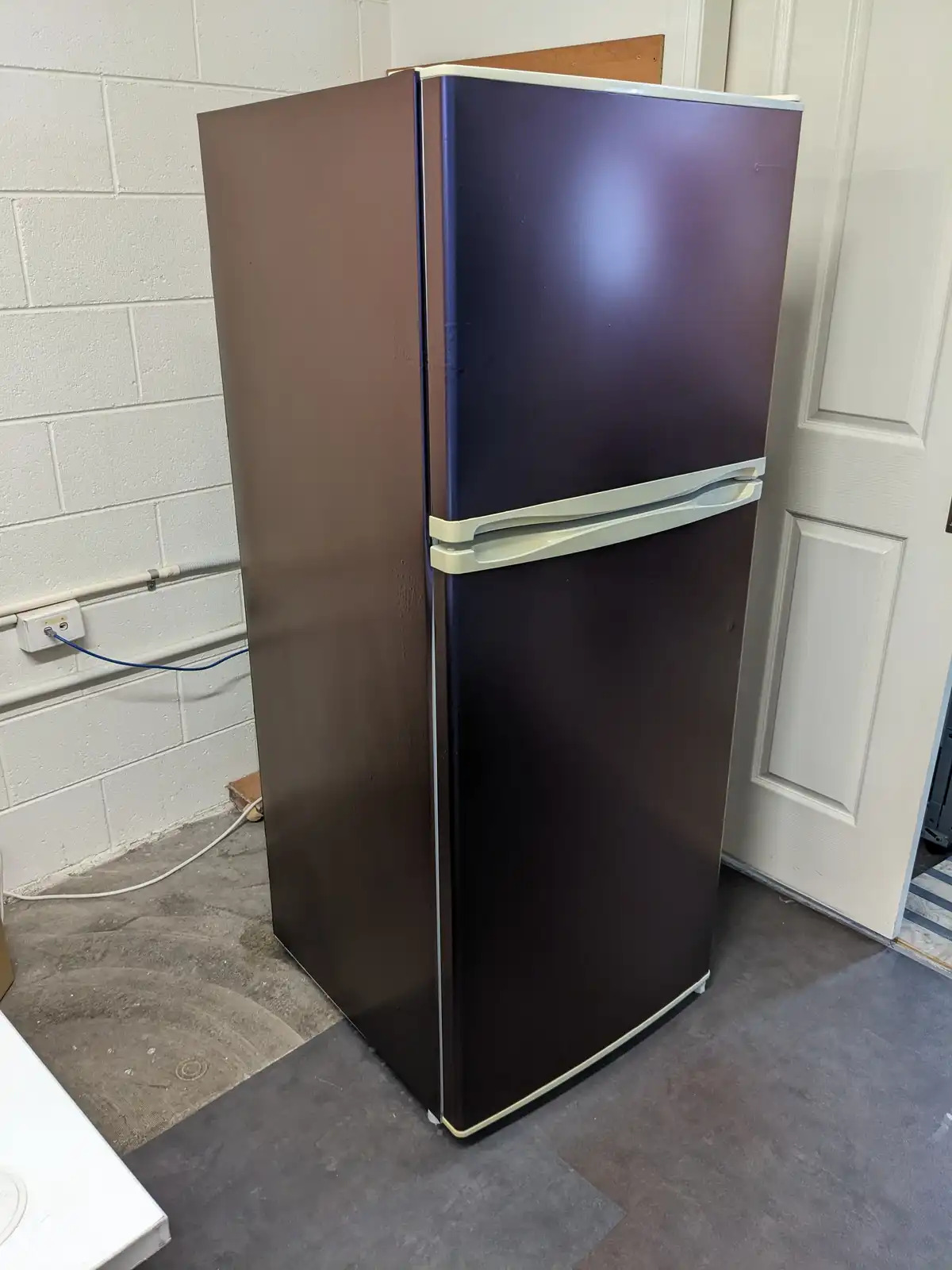 A two-door refrigerator with a dark brown, chocolate-like finish stands in a room with a white cinder block wall and a white…