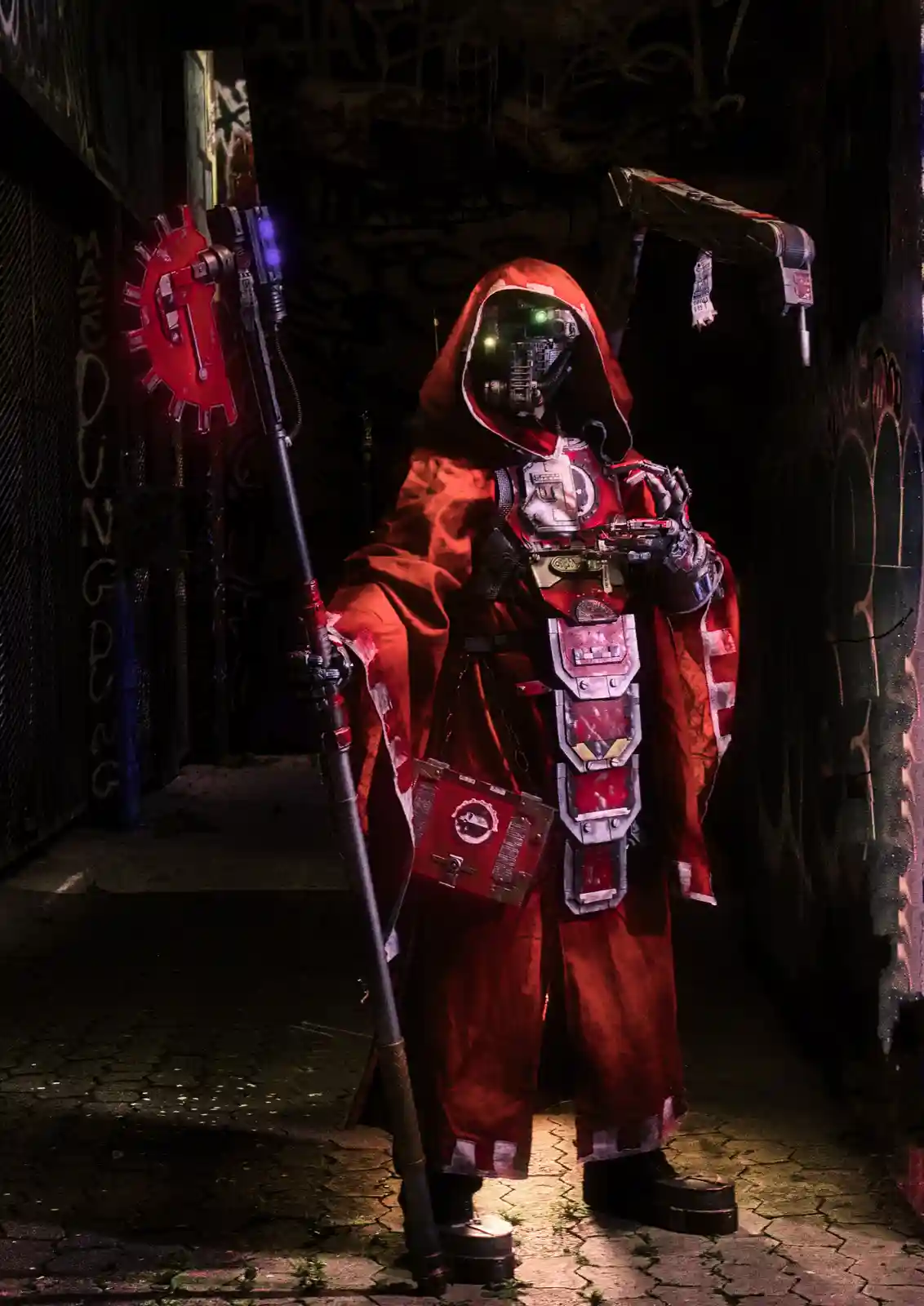 A person in a detailed red hooded costume with glowing green eyes and mechanical armor stands in a dark, graffiti-covered…