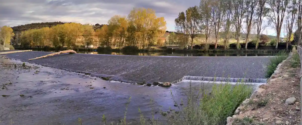 A wide river flows over a low weir, with autumn trees showing yellow and green leaves under a cloudy sky. The water cascades…