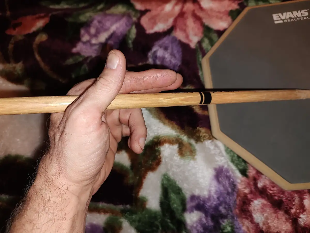 A close-up shot shows a person's hand holding a wooden drumstick horizontally, with the thumb and index finger gripping it…