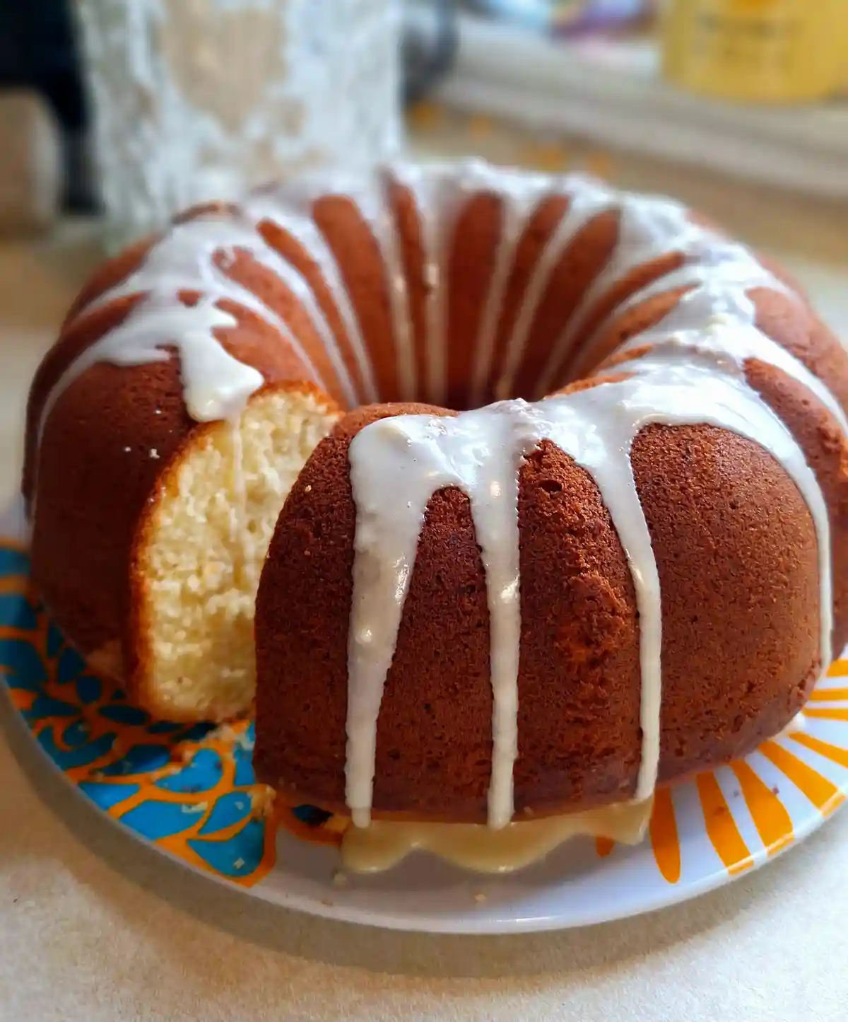 A close-up photo of a golden-brown bundt cake with white icing drizzled over it, sitting on a colorful plate. A slice has…
