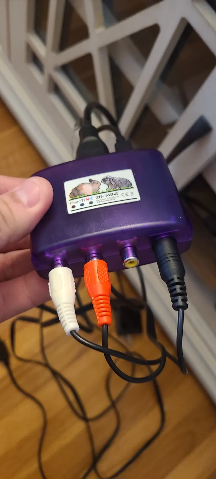 A hand holds a purple RetroTINK 2R-MINI device with multiple cables plugged in, including white, orange, and black…