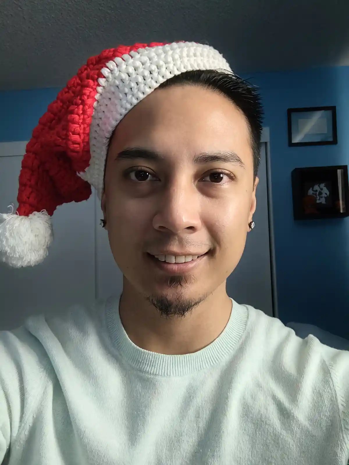A man with short dark hair and a light green sweater smiles at the camera while wearing a red and white crocheted Santa hat…