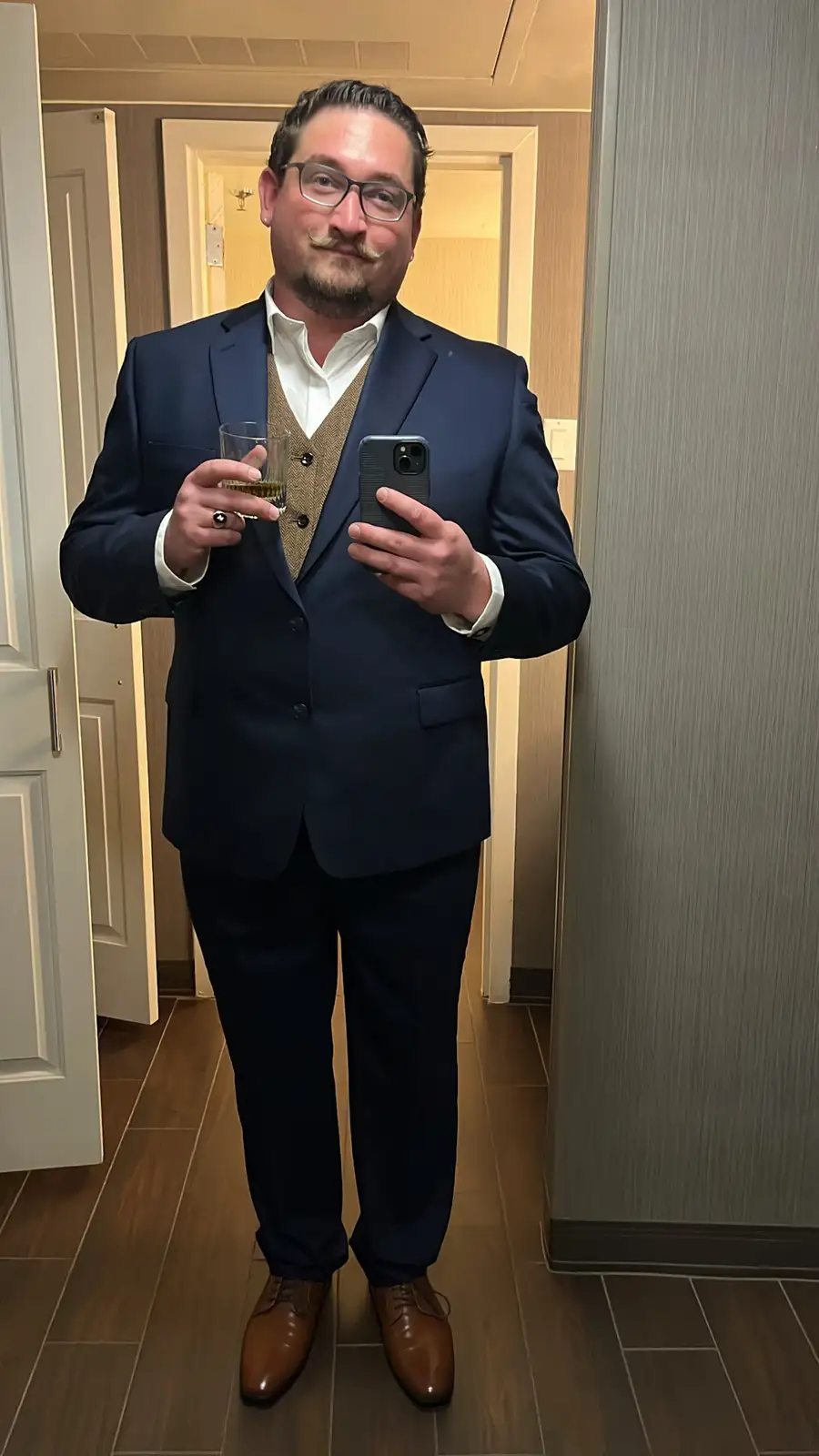 A man in a dark blue suit, white shirt, and brown vest takes a mirror selfie while holding a glass of whiskey in one hand…