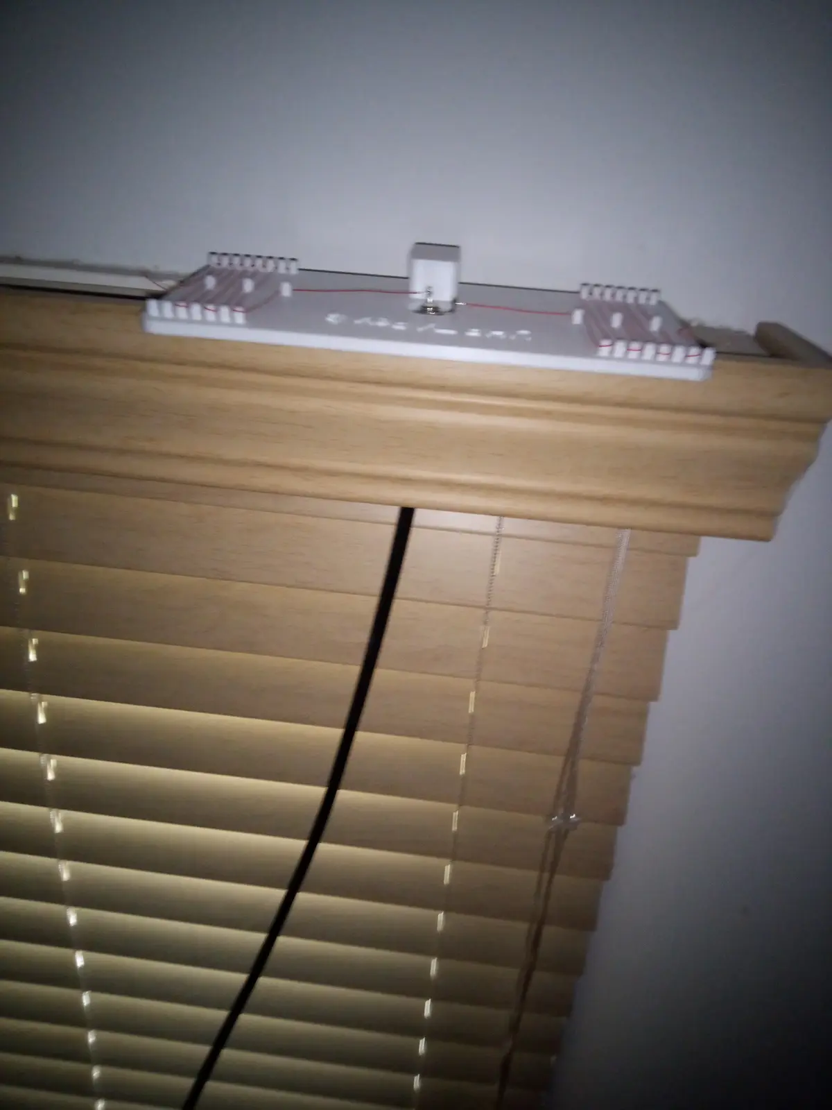 A white diy electronic circuit board with red wires and a small white box sits atop a wooden blind track, with light-colored…