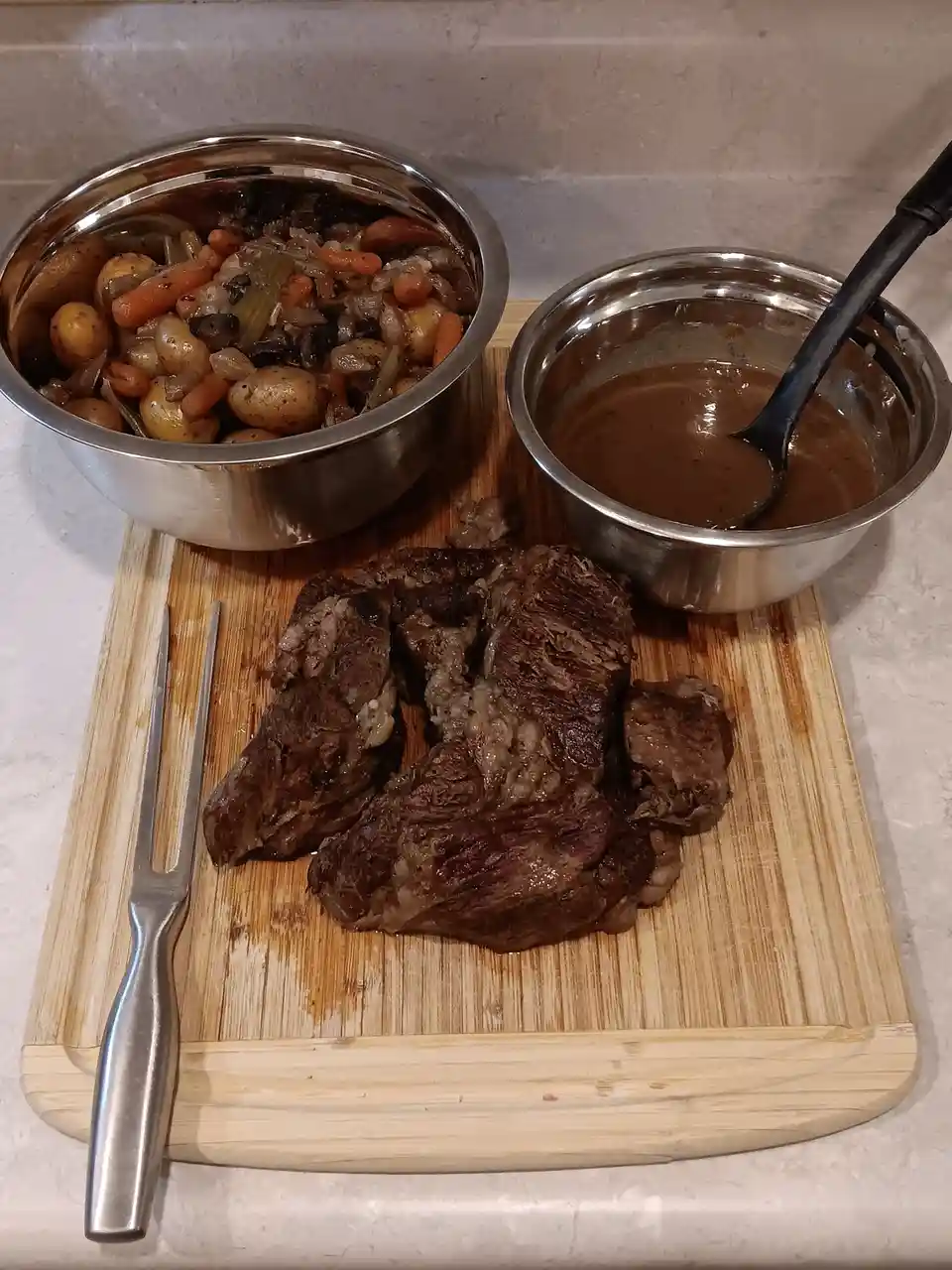 A wooden cutting board holds a large, shredded piece of cooked beef, a metal fork, and two stainless steel bowls. One bowl…