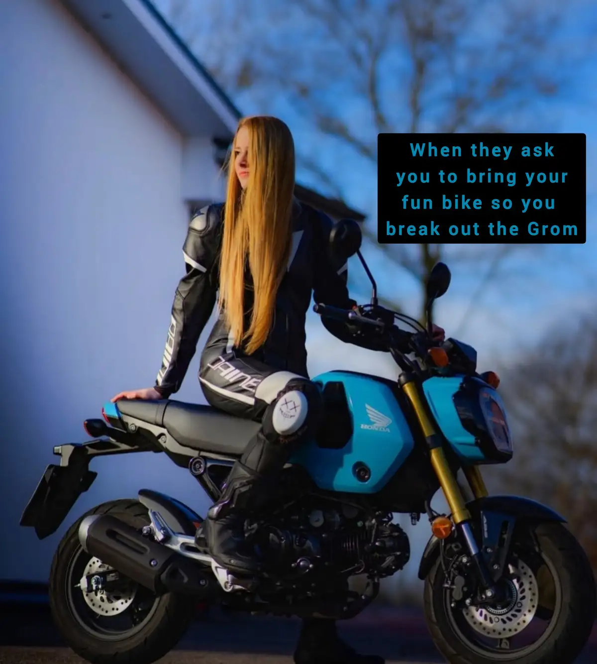 A woman with long blonde hair sits on a bright blue Honda Grom motorcycle, wearing full black and white motorcycle gear. The…