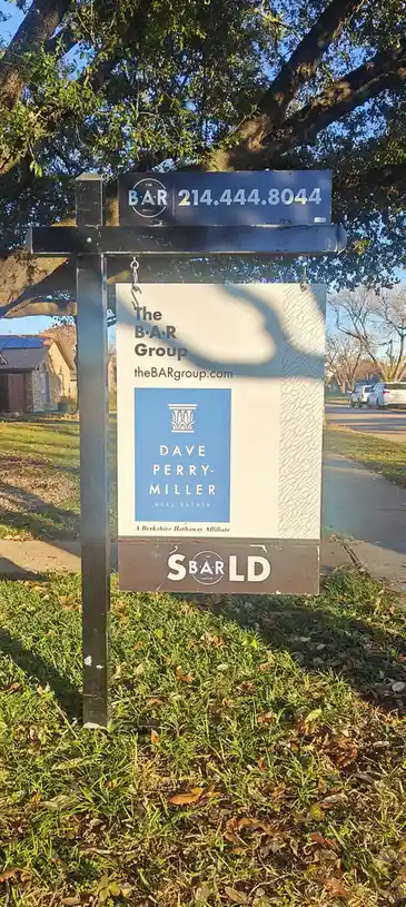 A real estate sign for 'The B·A·R Group' and 'Dave Perry-Miller Real Estate' stands in a grassy yard under a large tree. The…