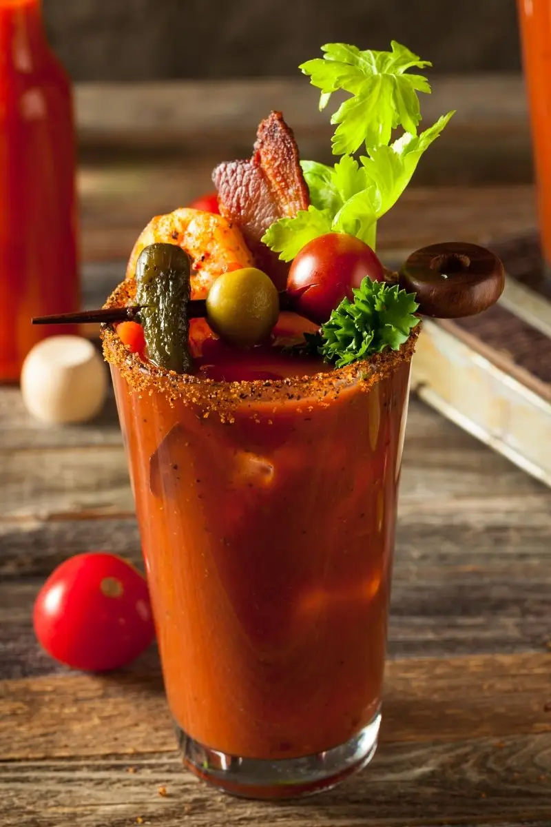 a tall glass filled with red tomato-based liquid, garnished with bacon, a pickle, olives, cherry tomatoes, celery leaves…