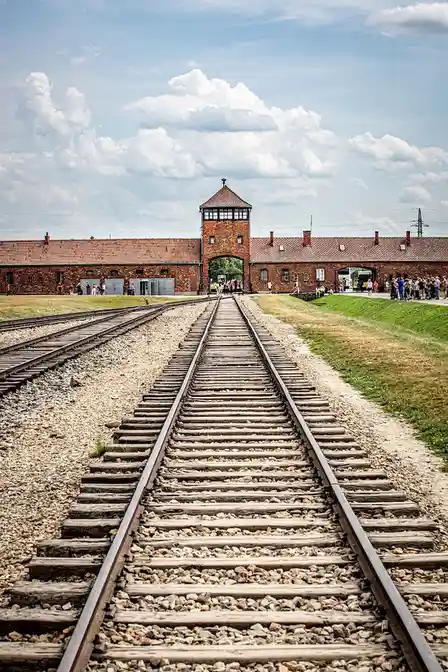 A photograph of the entrance to Auschwitz concentration camp, featuring railway tracks leading toward the iconic gate with…