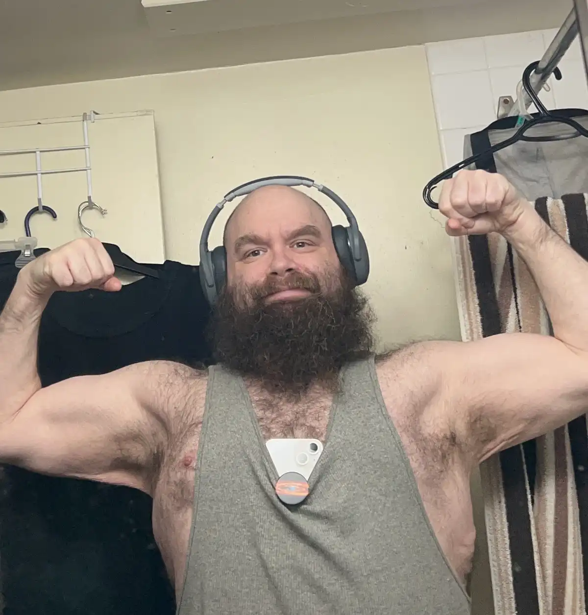 A bald man with a thick beard wearing grey over-ear headphones and a grey tank top flexes both arms while holding black…