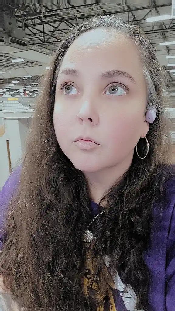 A young woman with long brown hair and a purple top looks upward with wide, unimpressed eyes in a large retail store. She…