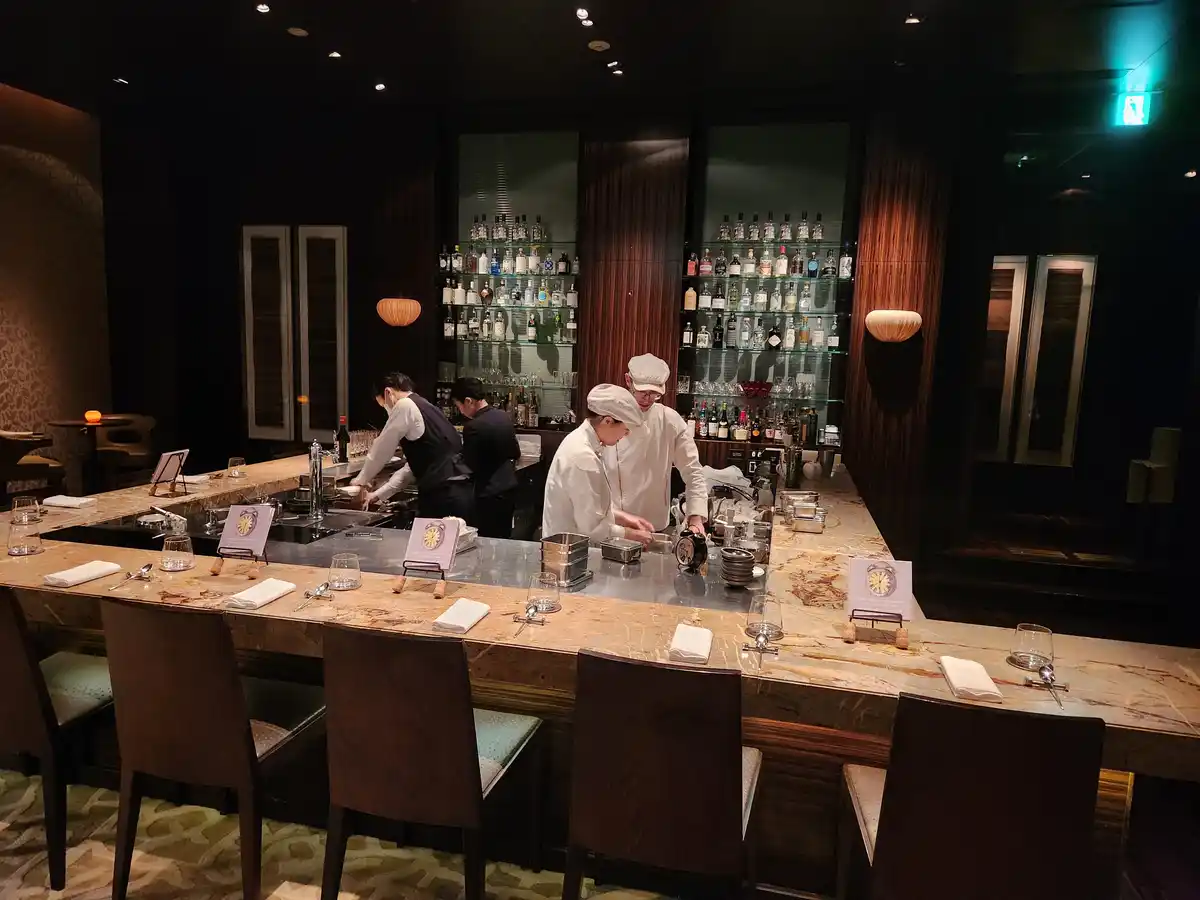 In a dimly lit, upscale bar with a long marble counter, several staff members in white chef hats and uniforms are preparing…