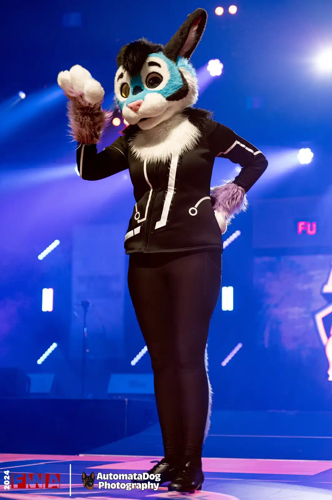 A person in a blue and black fursuit resembling a rabbit or fox stands on a stage under blue and pink lights, waving one paw…