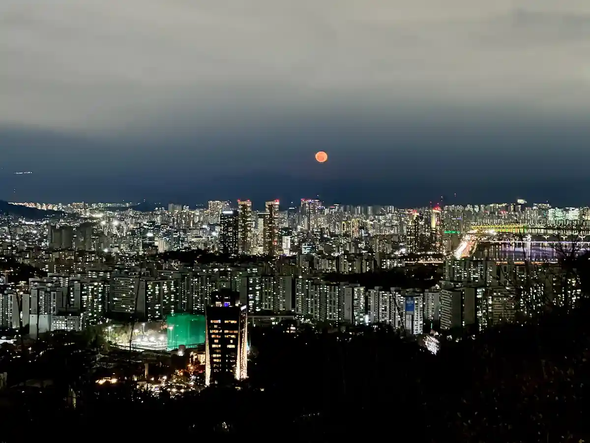 A nighttime cityscape photo shows a glowing orange moon above a densely lit urban skyline with numerous high-rise buildings…