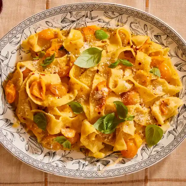 A top-down photo of a plate of pasta with orange tomatoes, basil leaves, and grated cheese, served on a decorative…