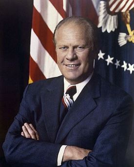 a formal portrait of a smiling man in a dark suit with arms crossed, standing in front of an american flag and a…