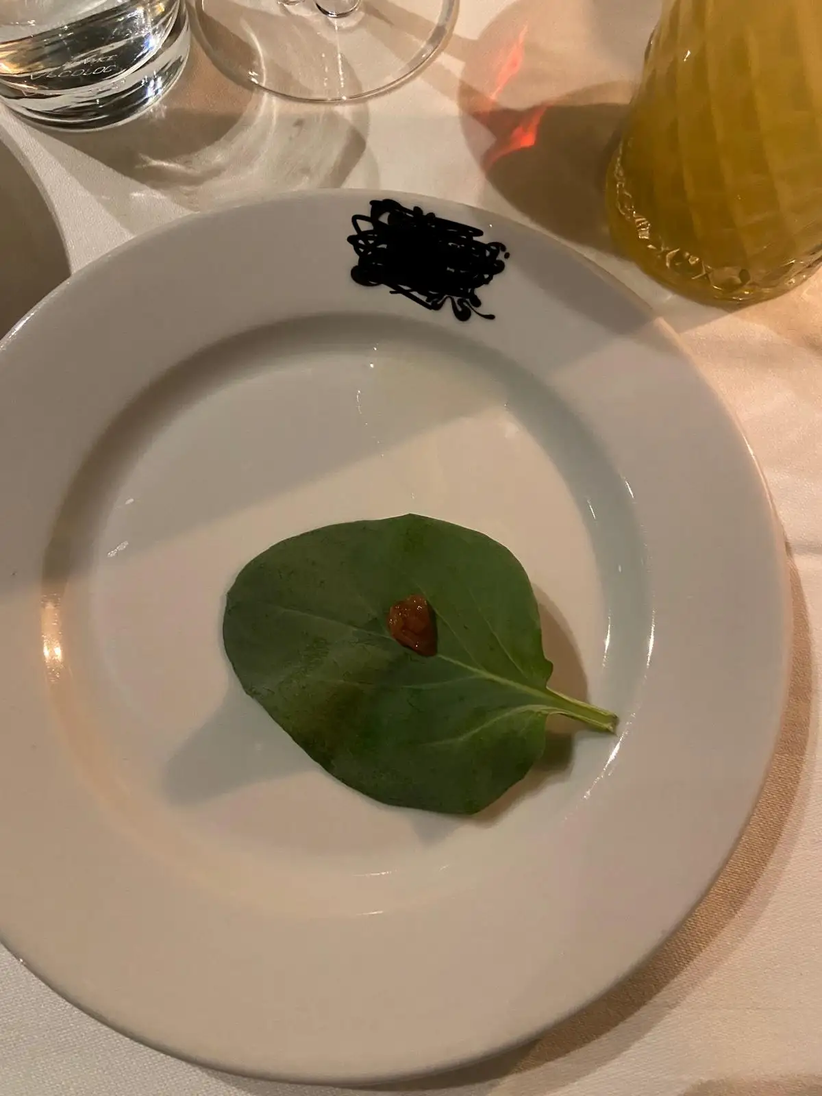 a minimalist restaurant dish featuring a single green leaf with a tiny dollop of orange sauce on it, placed on a white plate…