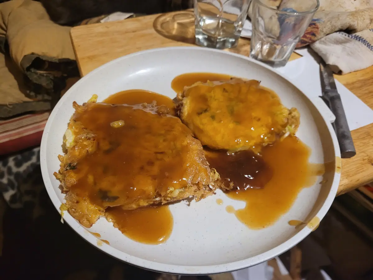 A white speckled plate holds two large, golden-brown fried food items, possibly sandwiches or patties, heavily drenched in a…